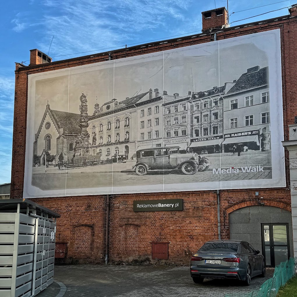 Duży baner reklamowy na ceglanej ścianie budynku przedstawiający czarno-białe zdjęcie zabytkowej zabudowy miejskiej z samochodem retro; widoczne zaparkowane auto osobowe i fragment otoczenia.