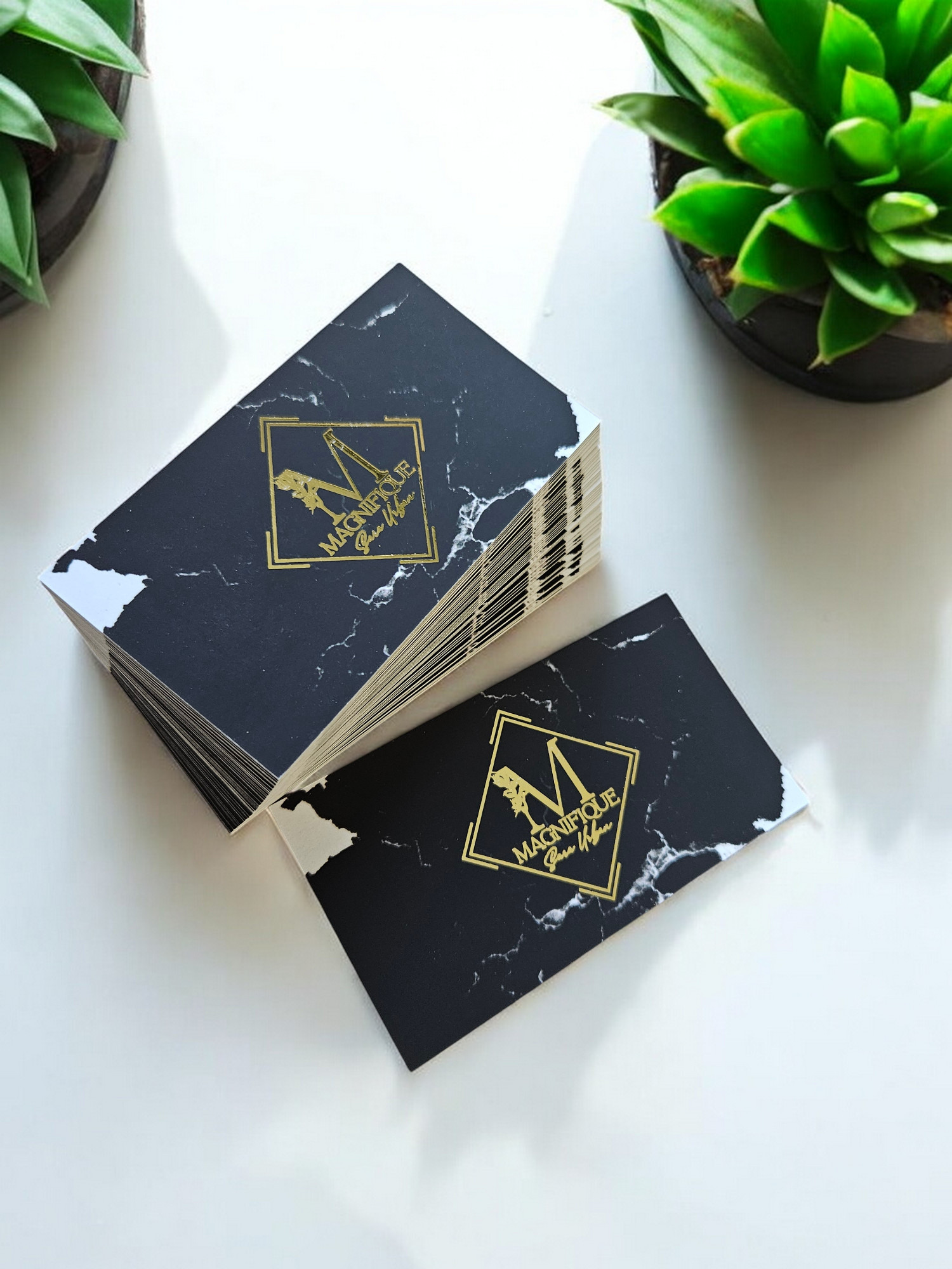 Stos czarnych, marmurkowych kart z złoconym logo 'Magnifique' na białym tle, obok doniczki z zieloną rośliną.