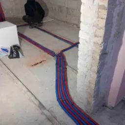 Instalacja ogrzewania podłogowego w trakcie realizacji: widoczne rury grzewcze w kolorze czerwonym i niebieskim, mocowane do podłoża, z fragmentem styropianu izolacyjnego i pracownikiem w tle.