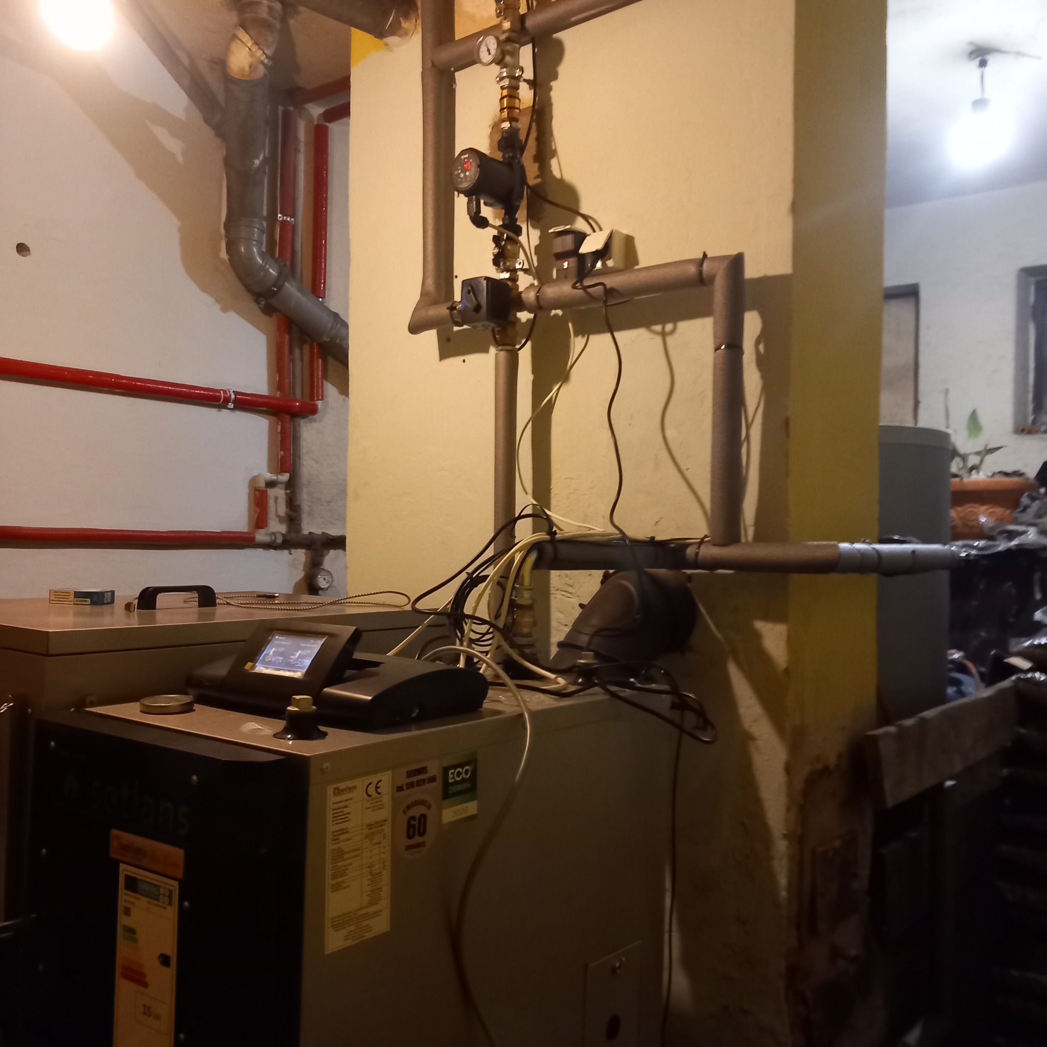 Nowoczesny piec centralnego ogrzewania marki Satlans z elektronicznym wyświetlaczem, podłączony do instalacji z rur miedzianych i pompy obiegowej w kotłowni.