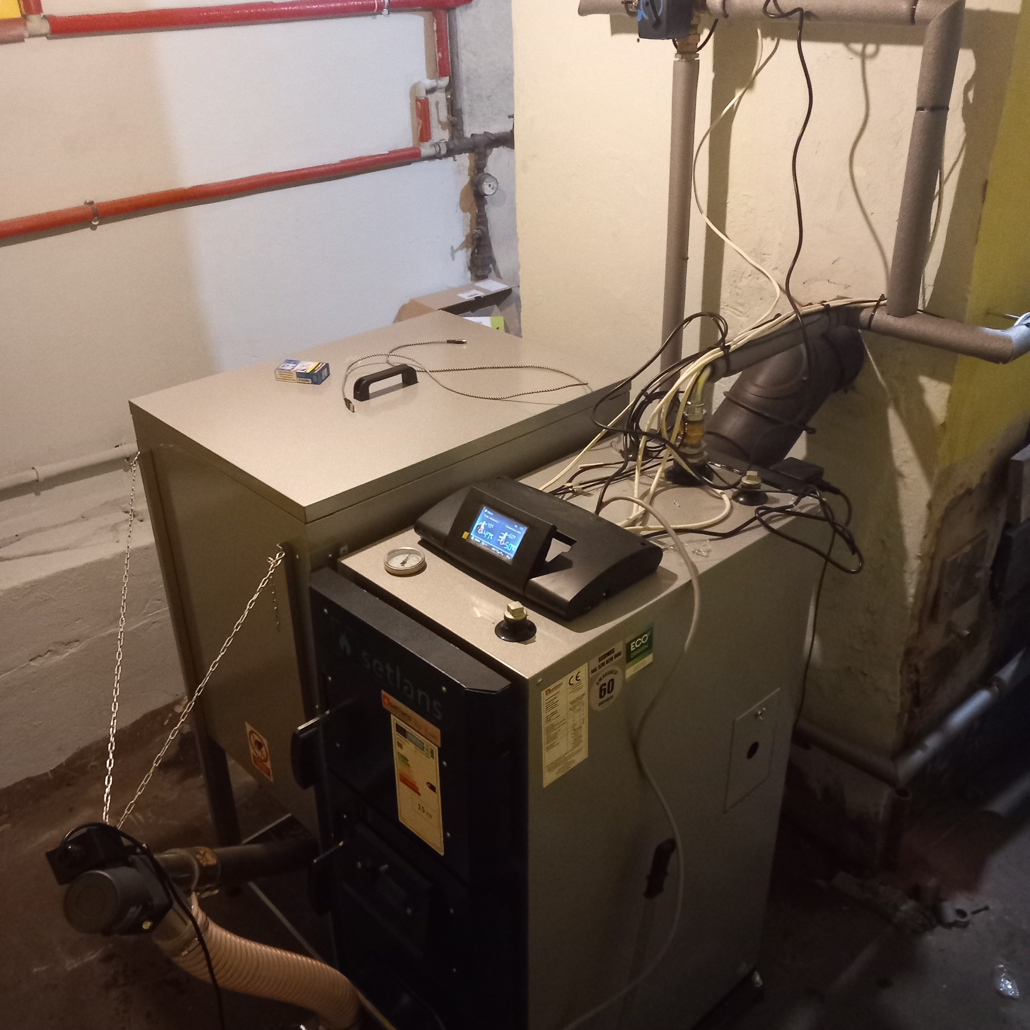 Nowoczesny piec na paliwo stałe marki Setlans z elektronicznym sterownikiem, zainstalowany w kotłowni z widocznymi rurami centralnego ogrzewania i elementami instalacji.