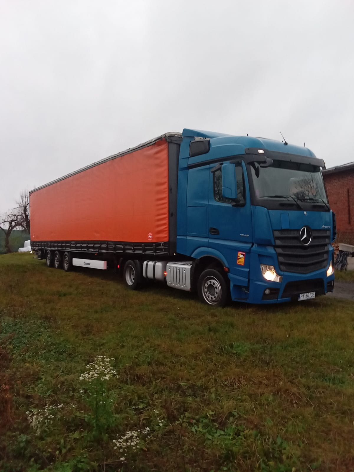 Niebieska ciężarówka Mercedes-Benz Actros z pomarańczową plandeką Krone zaparkowana na trawiastym terenie w pochmurny dzień.