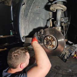 BAI-AUTO Oleksandr Baidiuk - Warsztat samochodowy: mężczyzna w rękawicach serwisuje tarczę hamulcową auta na podnośniku. Widoczne zawieszenie i błotnik. Podłoga warsztatu.