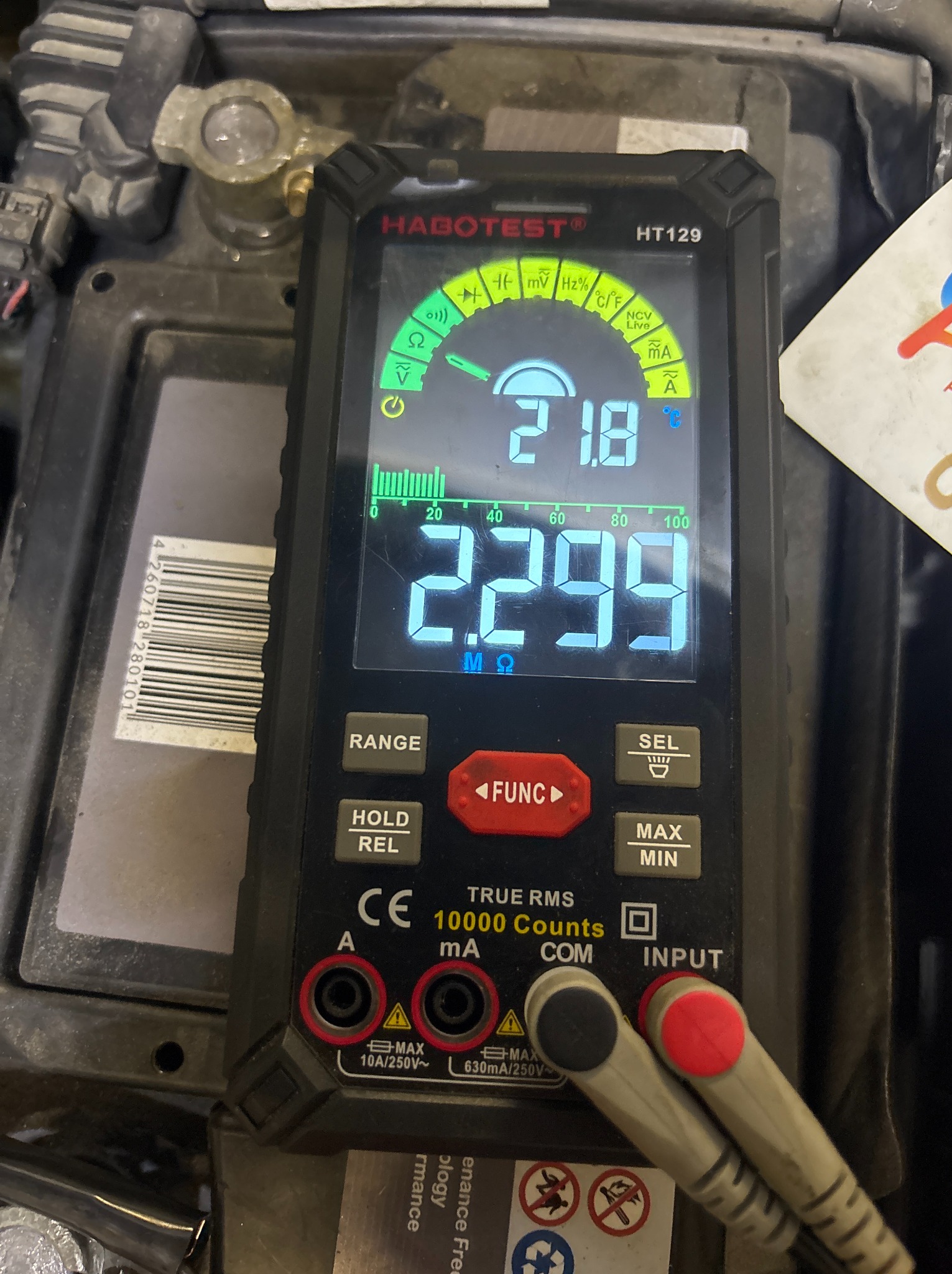 Pomiar napięcia akumulatora multimetrem Habotest HT129. Widoczne przewody pomiarowe i wyświetlacz z wartościami. Akumulator samochodowy w tle.