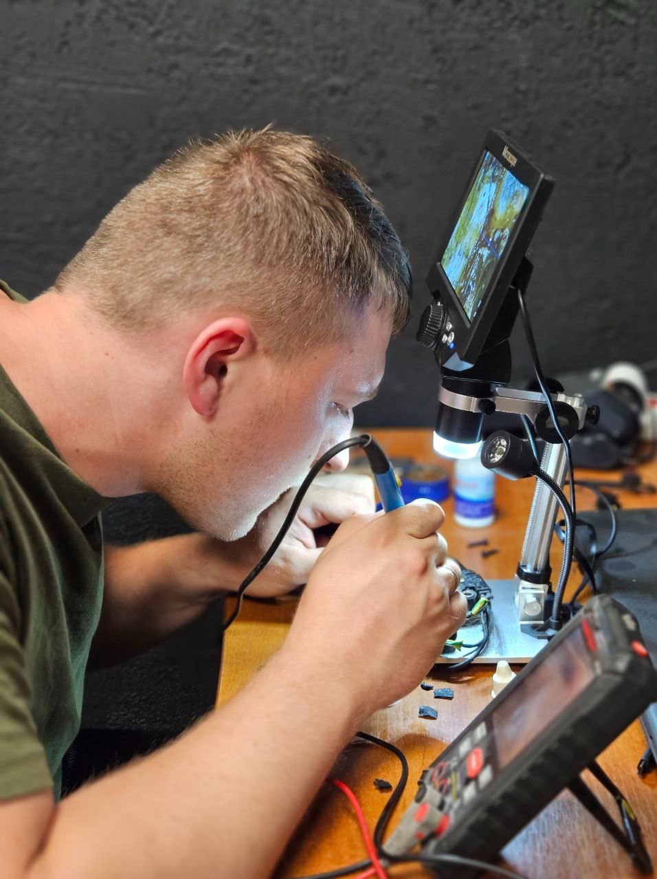 Elektryk precyzyjnie lutuje element elektroniczny pod mikroskopem z ekranem, używając lutownicy. Na stole widoczny multimetr i narzędzia.