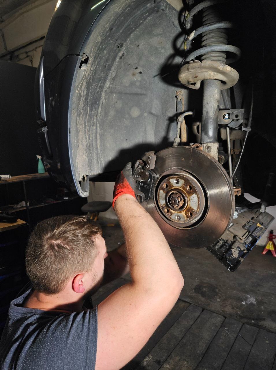 Warsztat samochodowy: mężczyzna w rękawicach serwisuje tarczę hamulcową auta na podnośniku. Widoczne zawieszenie i błotnik. Podłoga warsztatu.