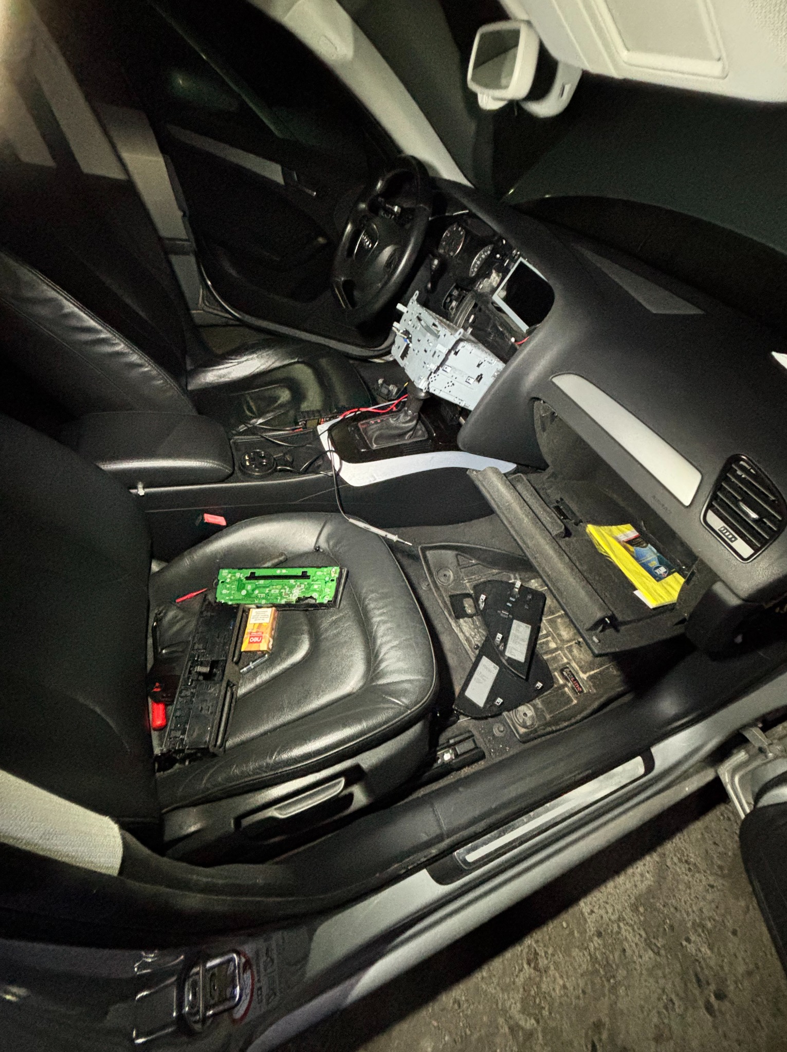 Wnętrze samochodu z rozmontowanym radiem i elementami elektrycznymi na skórzanym siedzeniu, wnętrze w trakcie naprawy, widoczne przewody i elementy elektroniki.