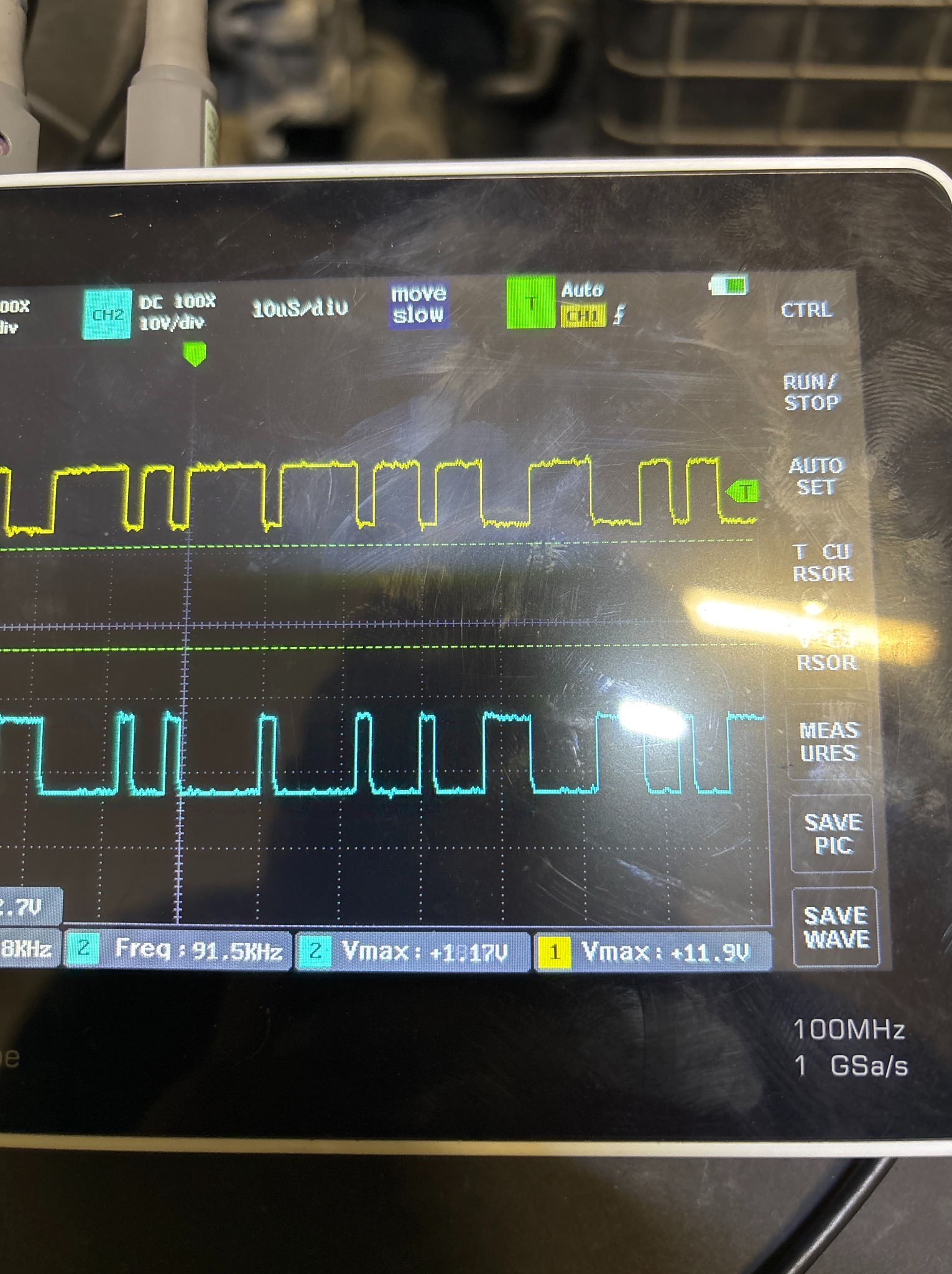 Oscyloskop z dwoma przebiegami sygnałów (żółty i niebieski) wyświetlającymi się na ekranie. Widoczne parametry: częstotliwość, napięcie.