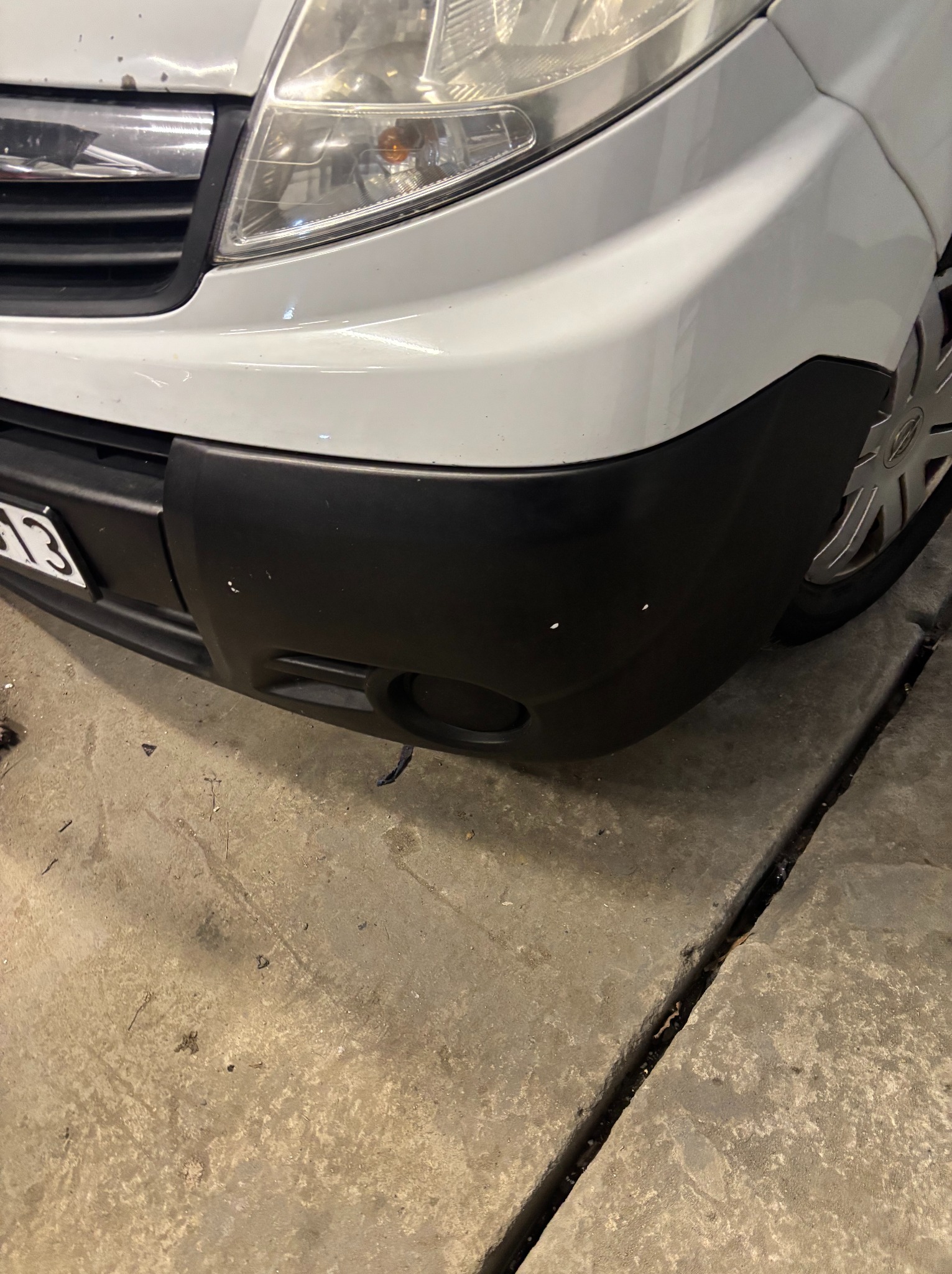 Fragment przedniego zderzaka białego samochodu z widocznymi drobnymi uszkodzeniami lakieru na czarnym elemencie. Widoczny fragment reflektora i koła.