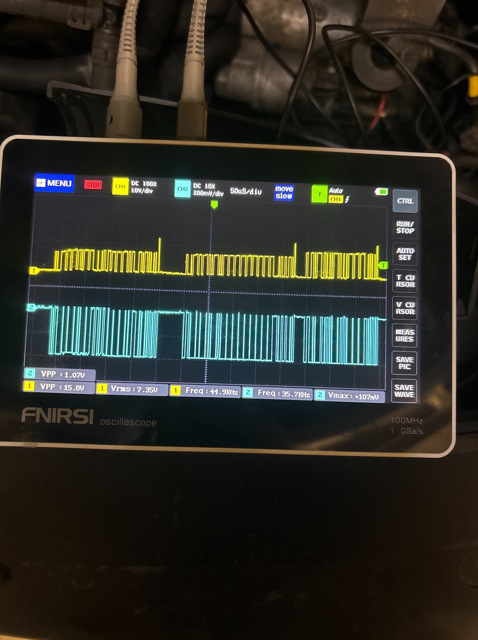 Oscyloskop FNIRSI z wyświetlonymi przebiegami sygnałów elektrycznych (żółty i niebieski) podczas diagnostyki samochodowej, widoczne kable pomiarowe.