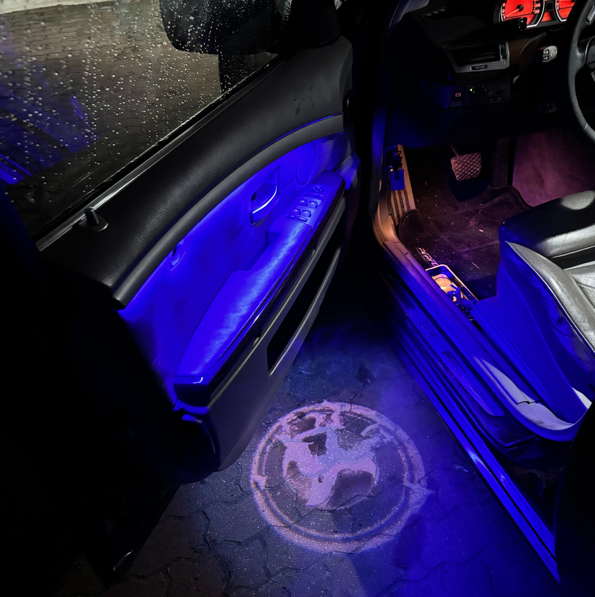 Nastrojowe oświetlenie wnętrza BMW w Warszawie, z podświetlanym logo marki na podłożu. Widoczny otwarty fragment drzwi i deska rozdzielcza.