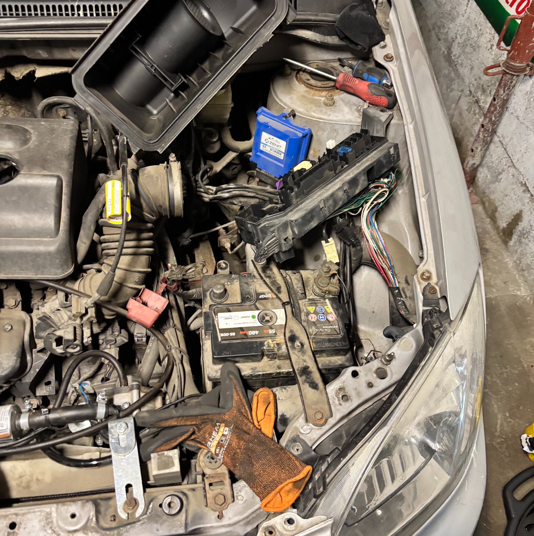 Otwarta maska srebrnego auta z widocznym silnikiem, akumulatorem i skrzynką bezpieczników. Na akumulatorze leżą robocze rękawice, obok widoczne narzędzia.