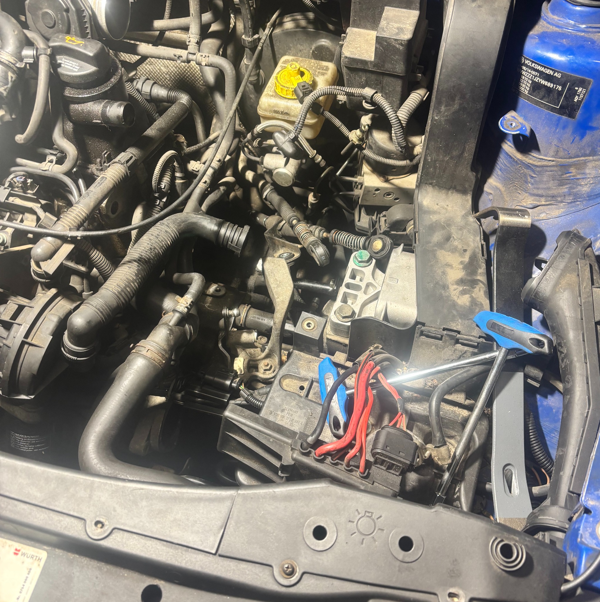 Otwarta maska niebieskiego samochodu, widoczny silnik w trakcie naprawy. Wokół silnika przewody, narzędzia i elementy mechaniczne.