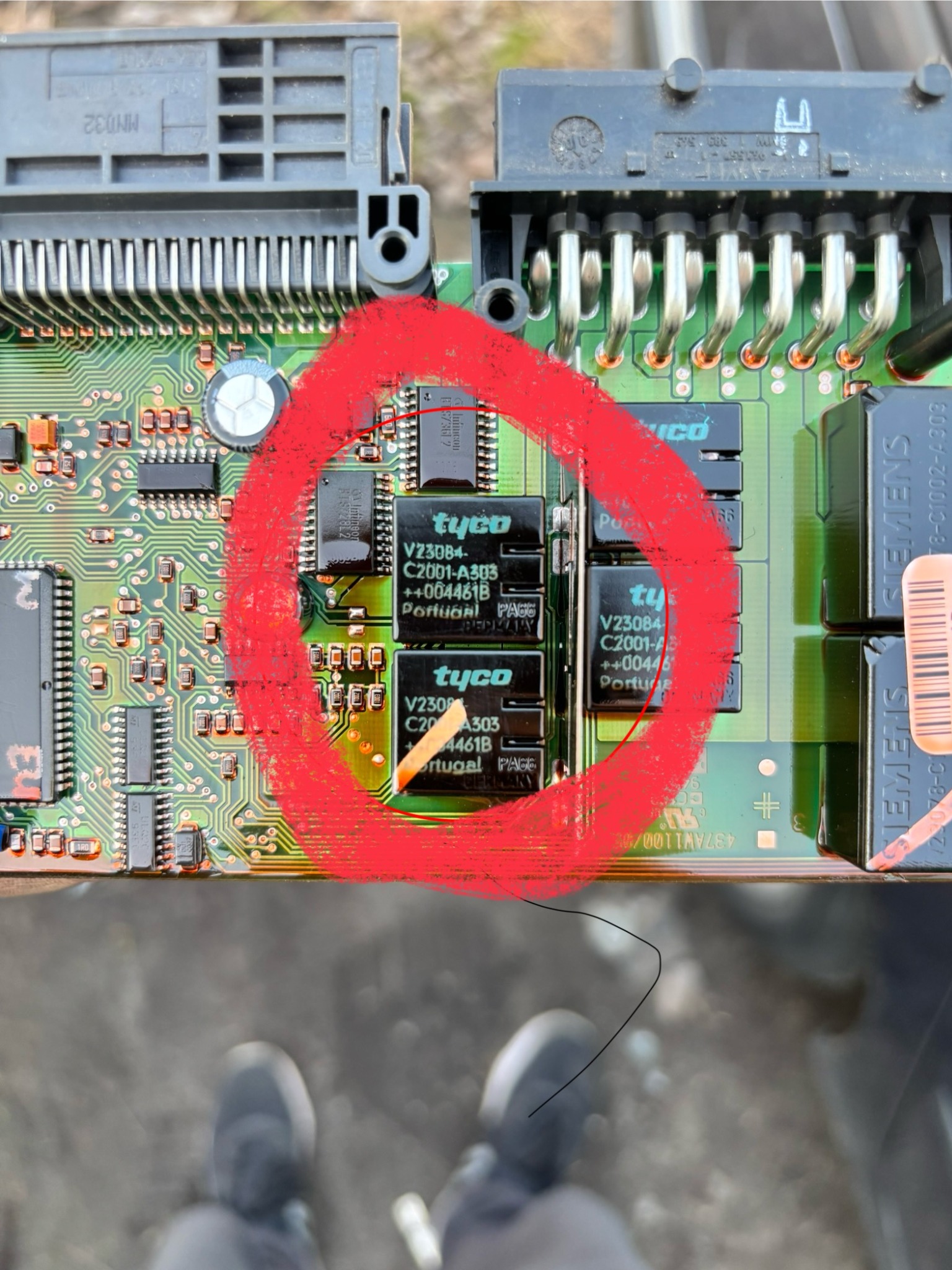 Płyta główna z elementami elektronicznymi, z zaznaczonym obszarem naprawy. Widoczne układy scalone Tyco V23084. Na dole kadru stopy osoby robiącej zdjęcie.