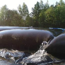 Przedsiębiorstwo Realizacyjne INORA Sp. z o.o. - Ujęcie z bliska na czarne, gumowe zbiorniki na wodę z wypływającą wodą, otoczone folią, w tle drzewa. Realizacja z zakresu ochrony środowiska.