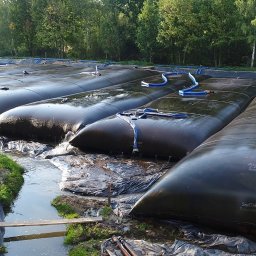 Przedsiębiorstwo Realizacyjne INORA Sp. z o.o. - Rząd wypełnionych worków geotekstylnych SoilTain DW z niebieskimi wężami na terenie z folią, otoczonych zielenią i drzewami, prawdopodobnie w trakcie procesu odwadniania.