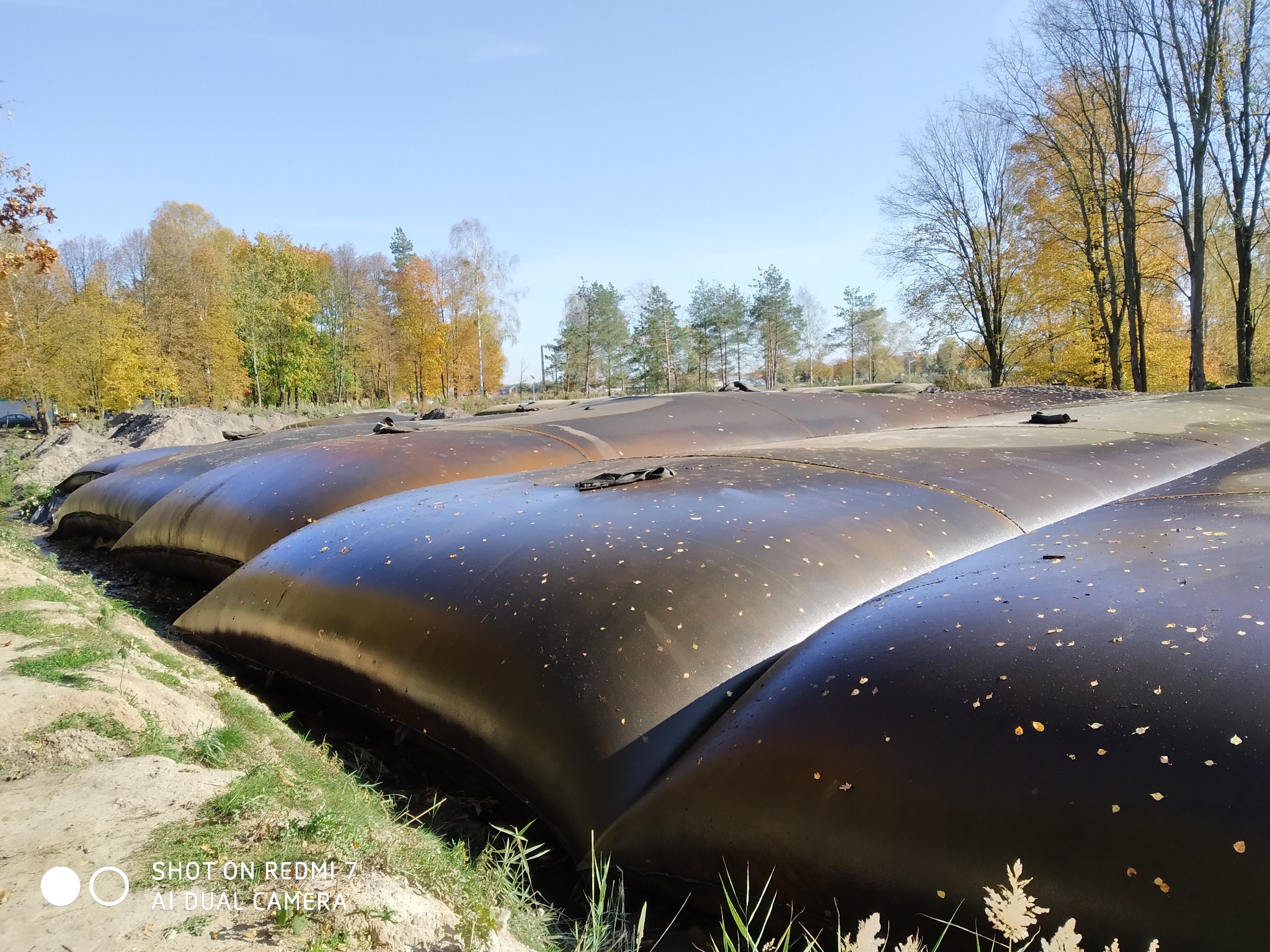 Rząd wypełnionych, czarnych worków geotekstylnych na brzegu wykopu, prawdopodobnie w celu stabilizacji gruntu. Jesienne drzewa w tle, błękitne niebo.