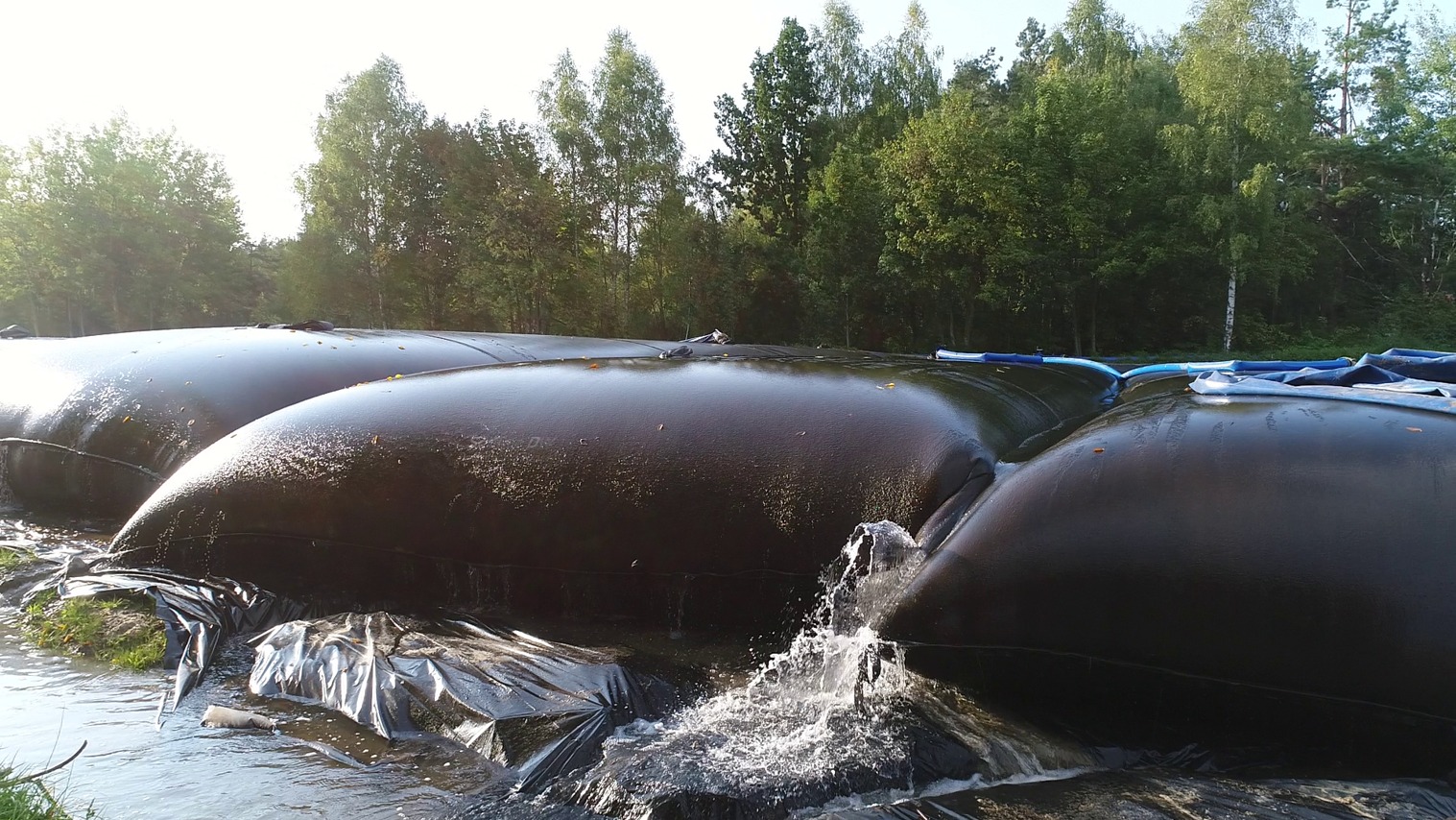 Ujęcie z bliska na czarne, gumowe zbiorniki na wodę z wypływającą wodą, otoczone folią, w tle drzewa. Realizacja z zakresu ochrony środowiska.