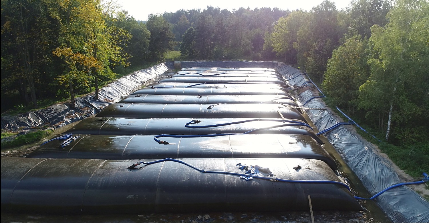 Długie, czarne zbiorniki na wodę w wykopanym stawie, zabezpieczone folią. Ujęcie z góry, widoczne drzewa i niebo z refleksami słońca na zbiornikach.