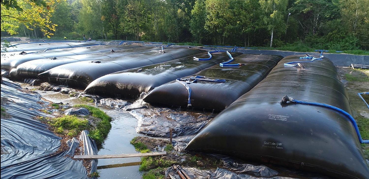 Rząd wypełnionych worków geotekstylnych SoilTain DW z niebieskimi wężami na terenie z folią, otoczonych zielenią i drzewami, prawdopodobnie w trakcie procesu odwadniania.