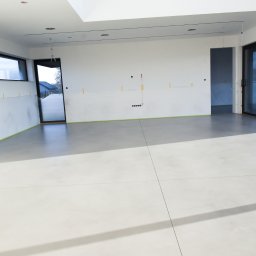 Pavistamp Polska Sławomir Potiopa - Jasnoszara posadzka w przestronnym wnętrzu z dużymi oknami i przygotowanymi ścianami do malowania, widoczne taśmy malarskie i folia ochronna.