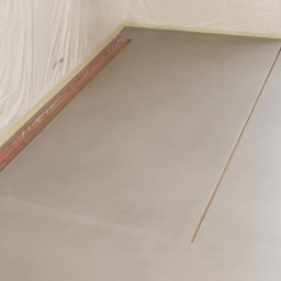 Pavistamp Polska Sławomir Potiopa - Świeżo wylana, jasnoszara posadzka w rogu pomieszczenia, zabezpieczona folią malarską i listwami przyściennymi, widoczna szczelina dylatacyjna.