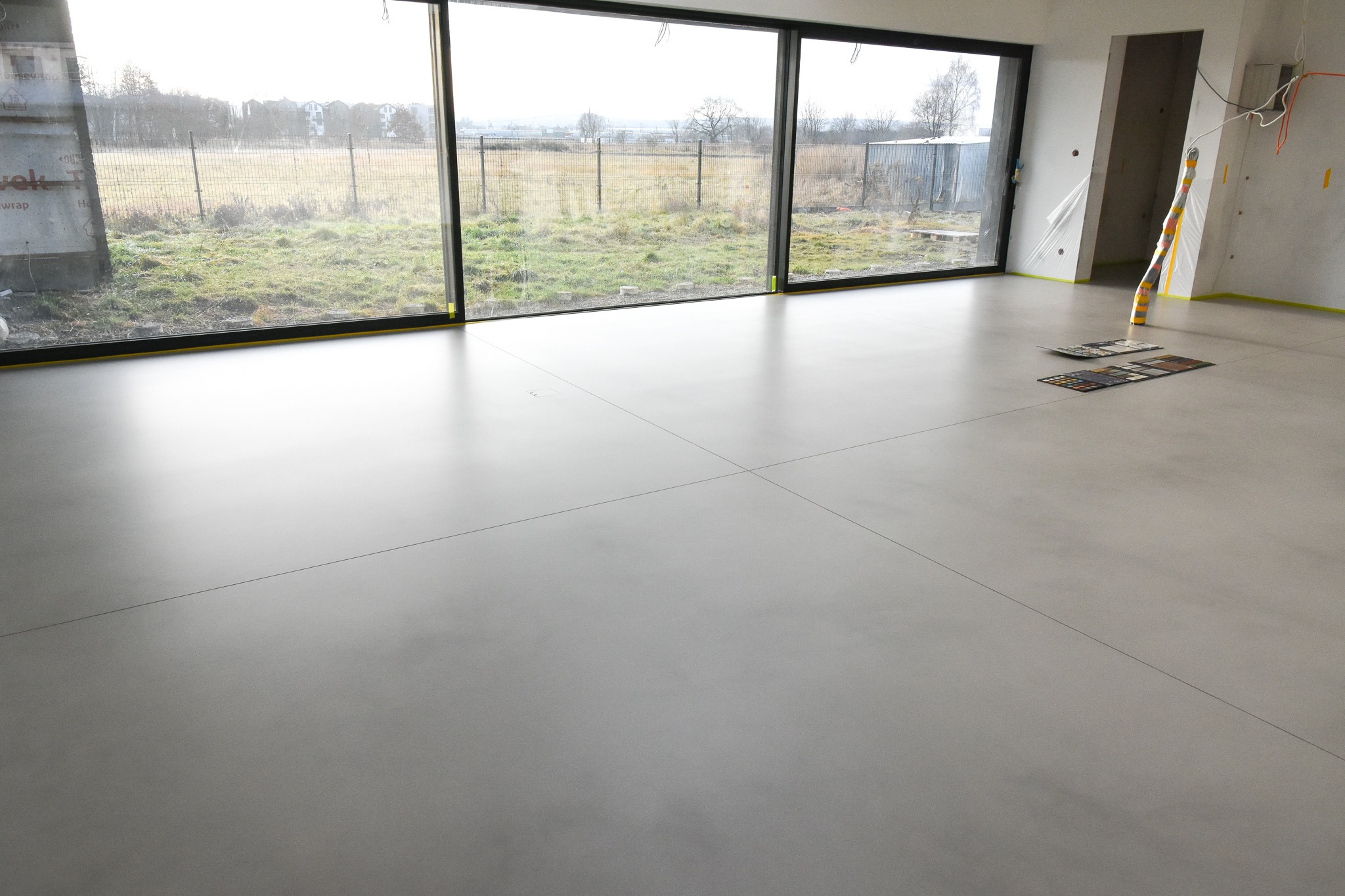 Szara, gładka posadzka w surowym wnętrzu z dużym oknem na ogród, próbki kolorów pod ścianą.