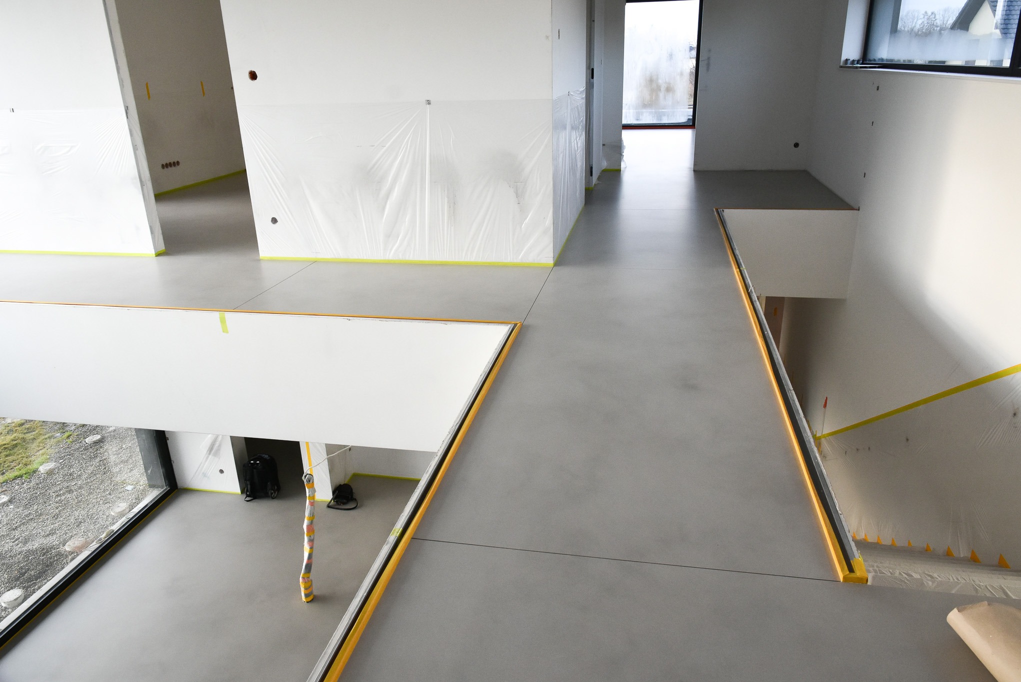 Nowoczesne wnętrze z szarą posadzką mikrocementową w trakcie prac wykończeniowych; ściany zabezpieczone folią i taśmą malarską, widok z góry na dwóch poziomach.