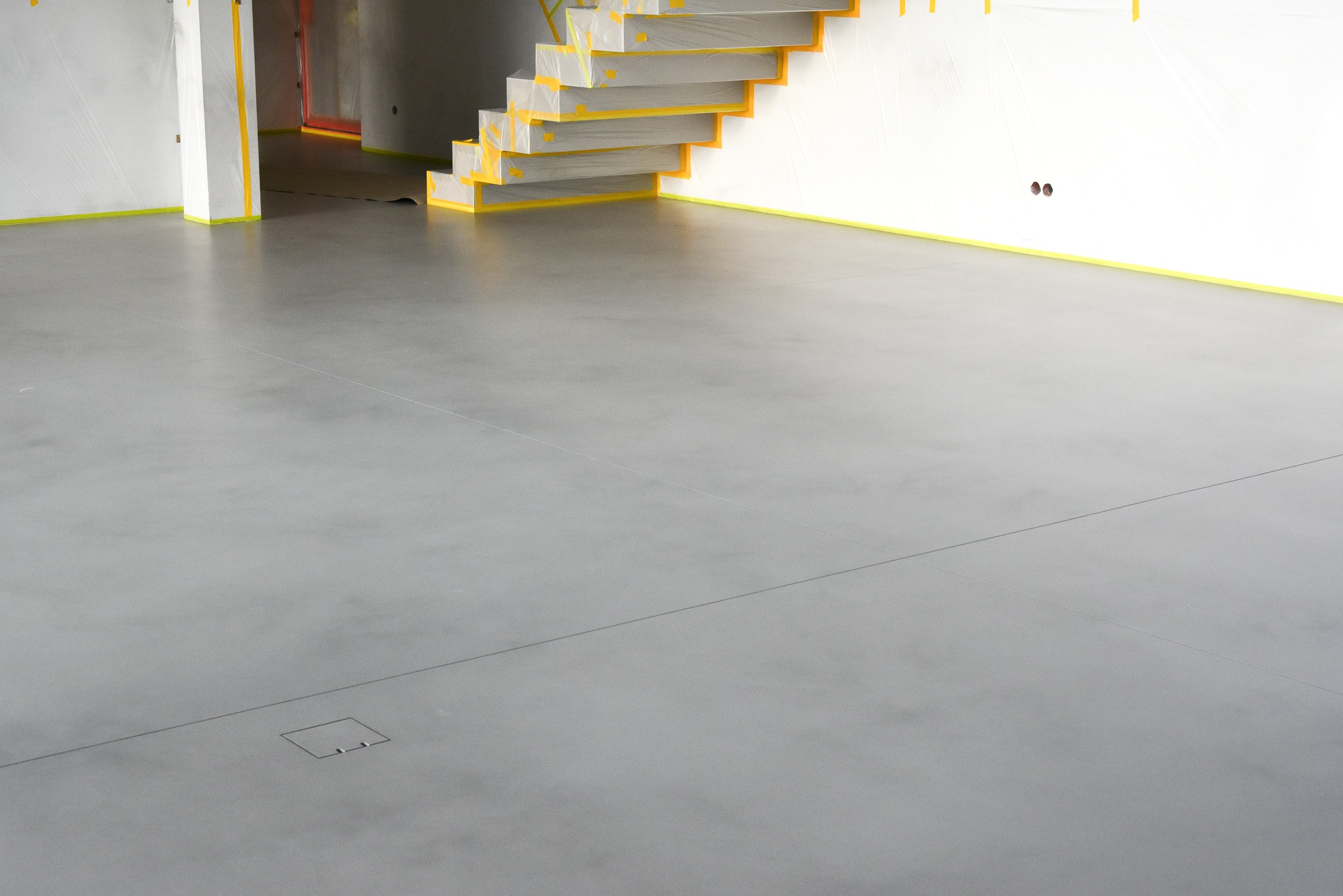Szara posadzka w surowym wnętrzu, zabezpieczone schody i ściany taśmą malarską, widoczna klapa rewizyjna w podłodze.