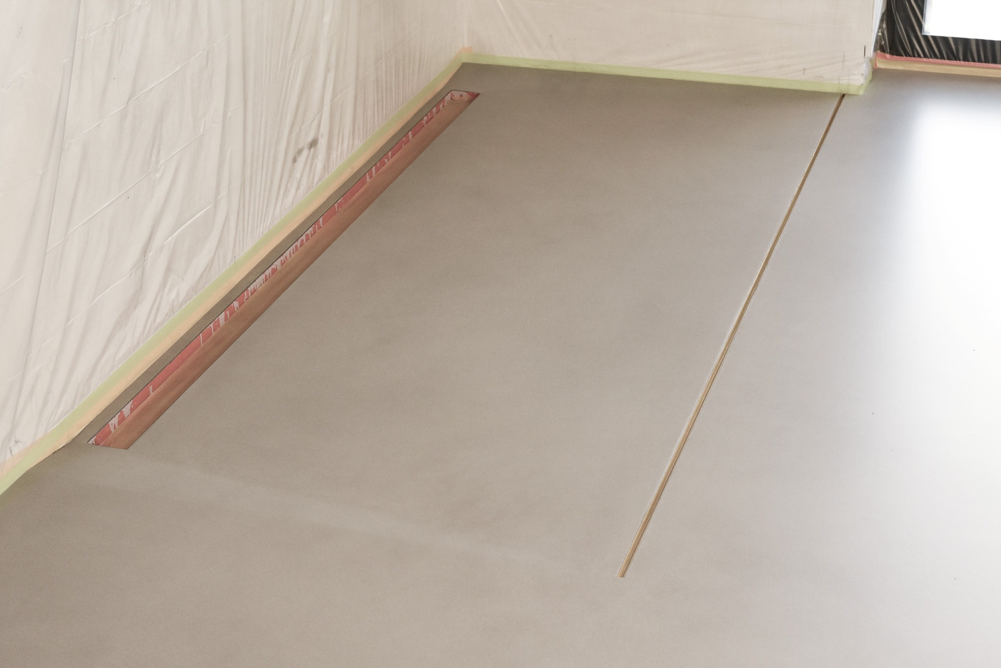 Świeżo wylana, jasnoszara posadzka w rogu pomieszczenia, zabezpieczona folią malarską i listwami przyściennymi, widoczna szczelina dylatacyjna.