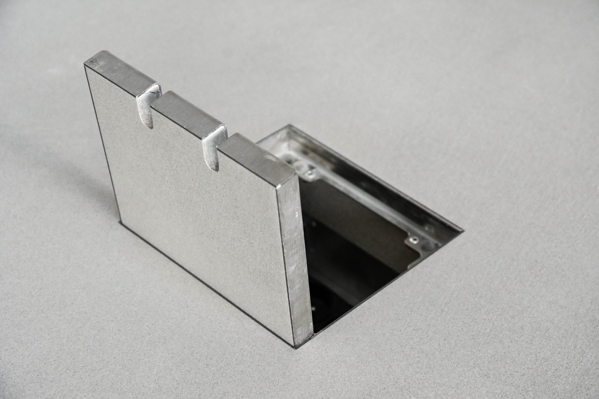 Szczegółowe ujęcie metalowej obudowy wpuszczonej w szarą posadzkę z mikrocementu, widoczne wycięcia na kable.