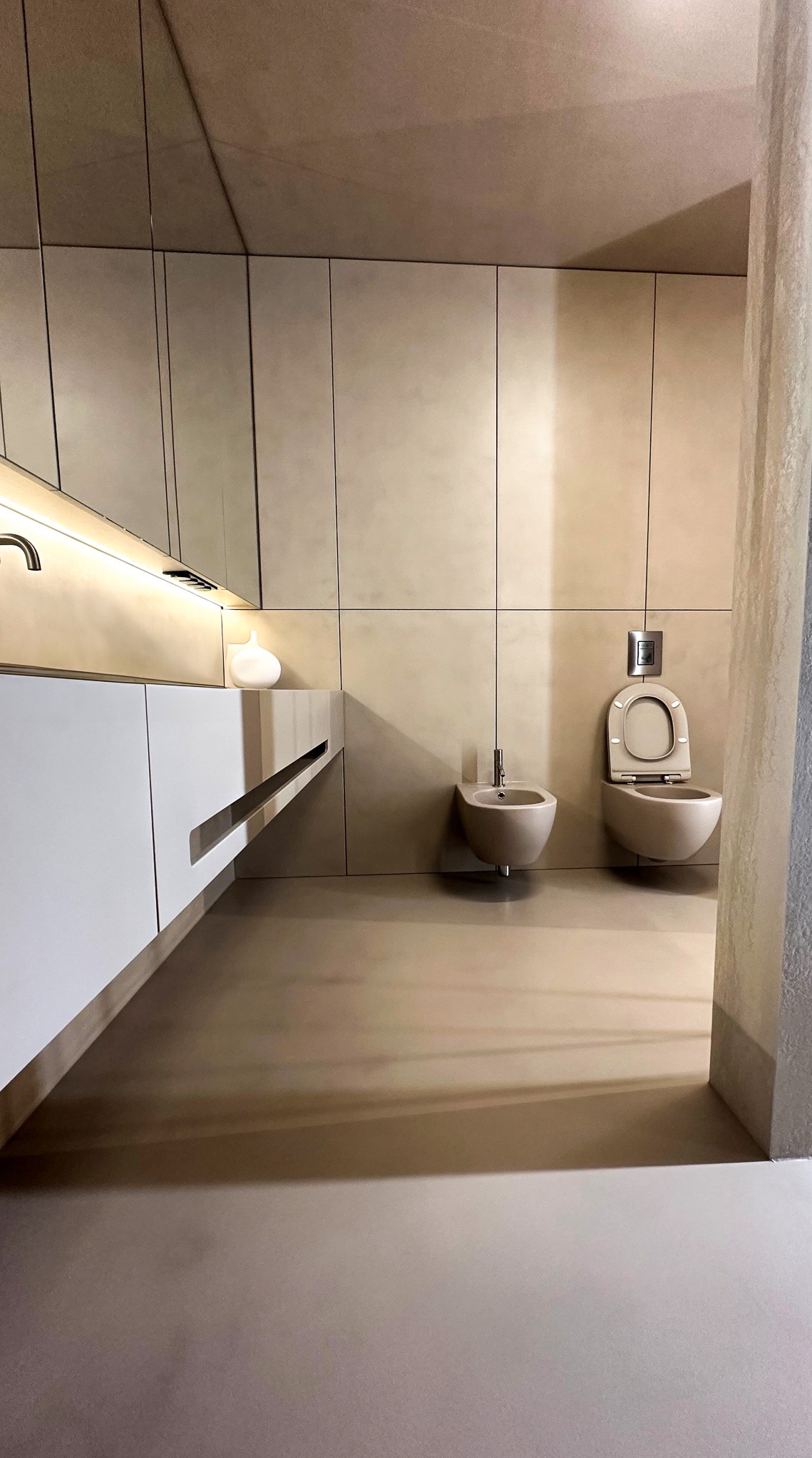 Minimalistyczna łazienka w odcieniach beżu z toaletą, bidetem i umywalką z szafką, wykończona jednolitą powierzchnią podłogi i ścian.