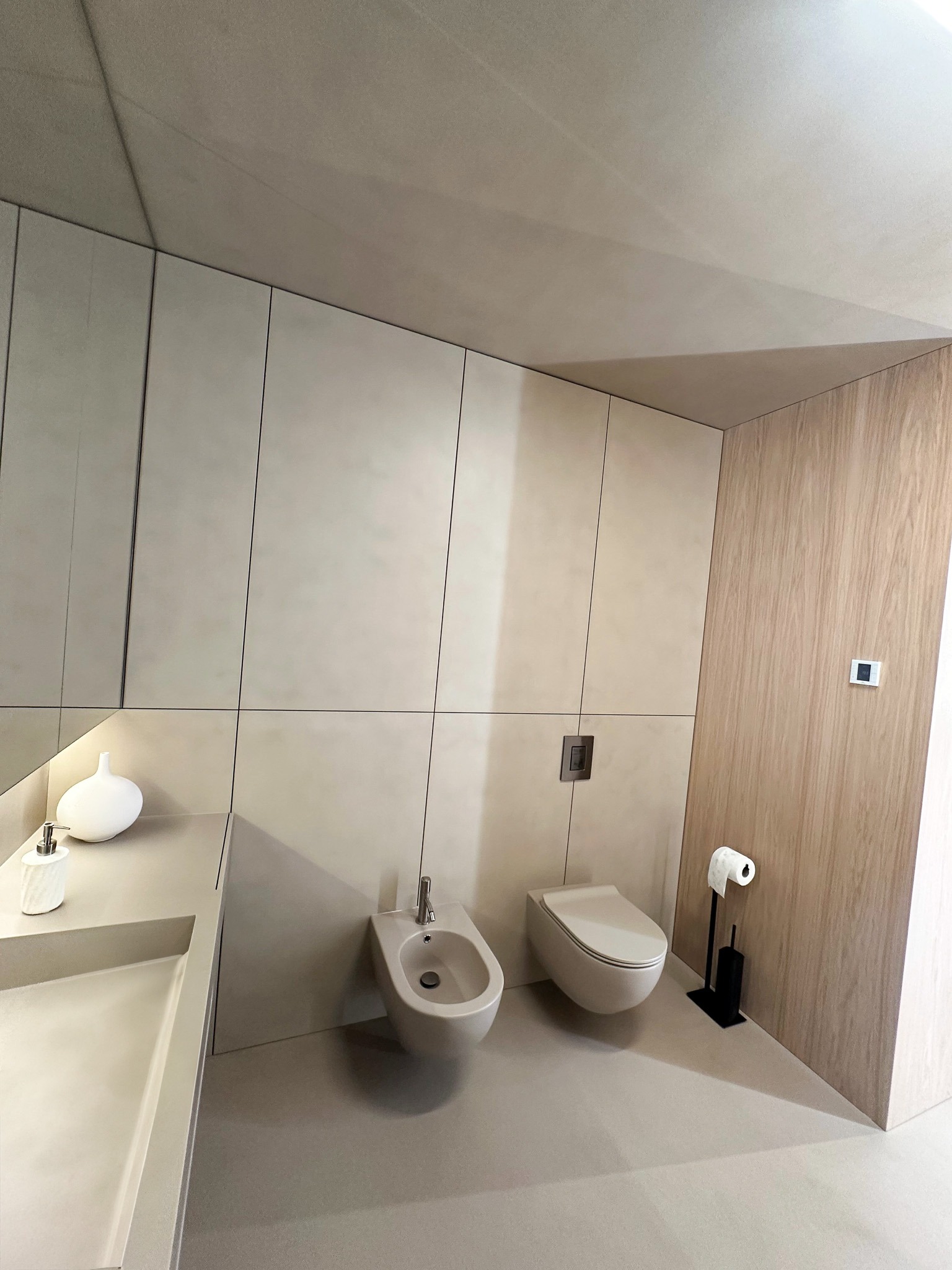 Minimalistyczna łazienka z beżową, jednolitą posadzką i ścianami, wisząca toaleta i bidet, umywalka zintegrowana z blatem, drewniana okładzina ściany z włącznikiem.