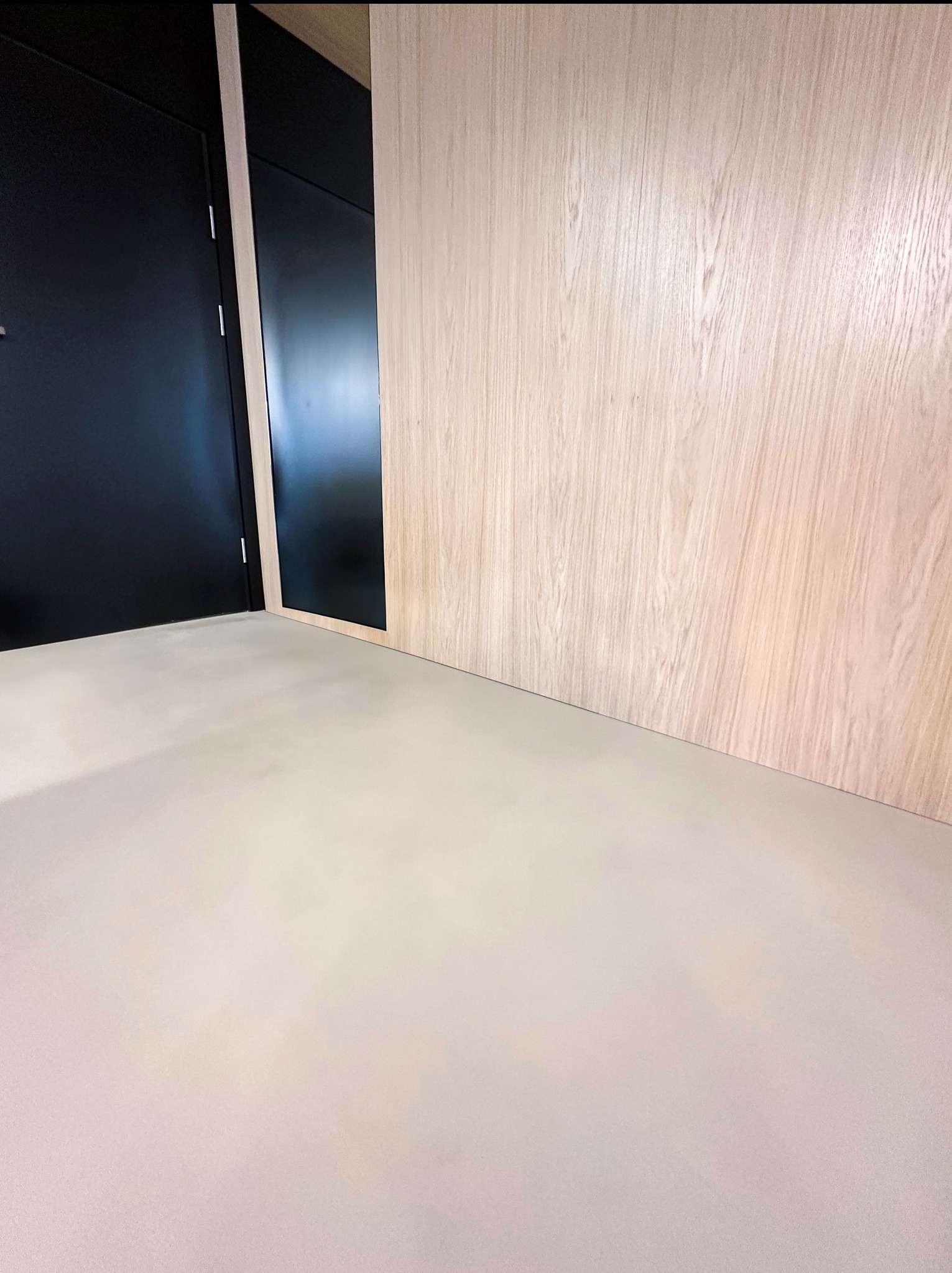 Jasnoszara, gładka posadzka w minimalistycznym wnętrzu z czarnymi drzwiami i drewnianą okładziną ściany.