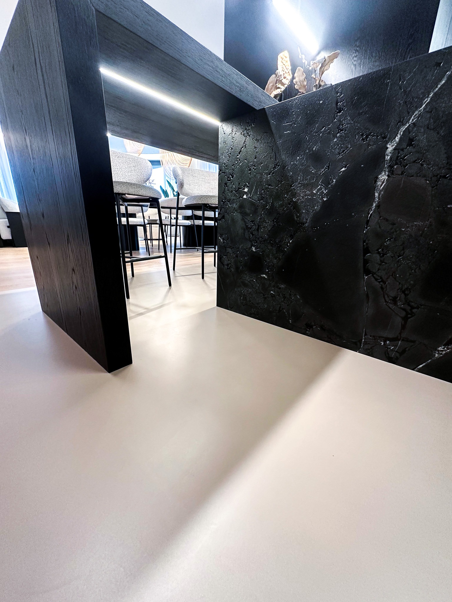 Jasna, jednolita posadzka w nowoczesnej kuchni z wyspą wykończoną czarnym marmurem i drewnianymi elementami, widoczne krzesła barowe w tle.