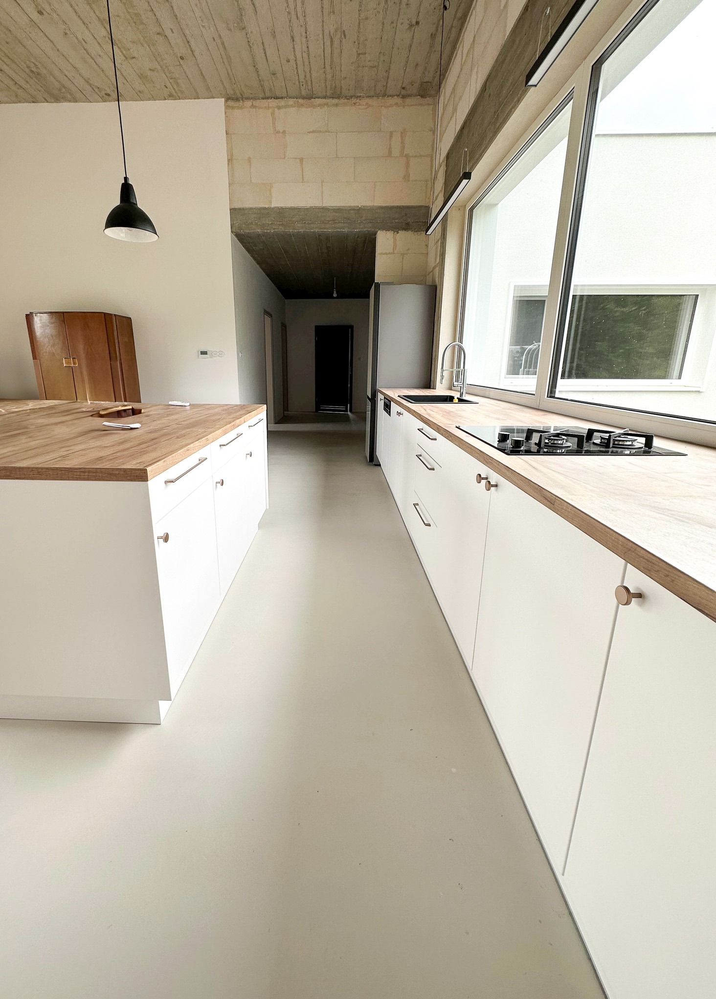 Minimalistyczna kuchnia z białymi szafkami i drewnianym blatem, posadzka z mikrocementu w odcieniach szarości, widoczny surowy betonowy strop i wisząca lampa.