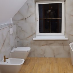 Mg Bud - Jasna łazienka na poddaszu z marmurowymi płytkami ściennymi i drewnopodobną podłogą. Uwagę przykuwa złota armatura przy bidecie i odbicie osoby w oknie.