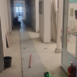 Mg Bud - Kładzenie szarych płytek w długim korytarzu, poziomowanie laserem krzyżowym. Widoczny kubeł, poziomica i zapasowe płytki.