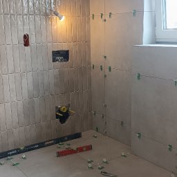 Mg Bud - Układanie beżowych płytek w łazience z użyciem poziomicy i klipsów. Widoczne instalacje i okno. Praca w toku, oświetlenie halogenowe.
