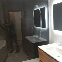 Mg Bud - Nowoczesna łazienka z czarnymi płytkami, szklaną kabiną prysznicową i drewnianą szafką pod umywalką. Minimalistyczny design z oświetleniem LED.