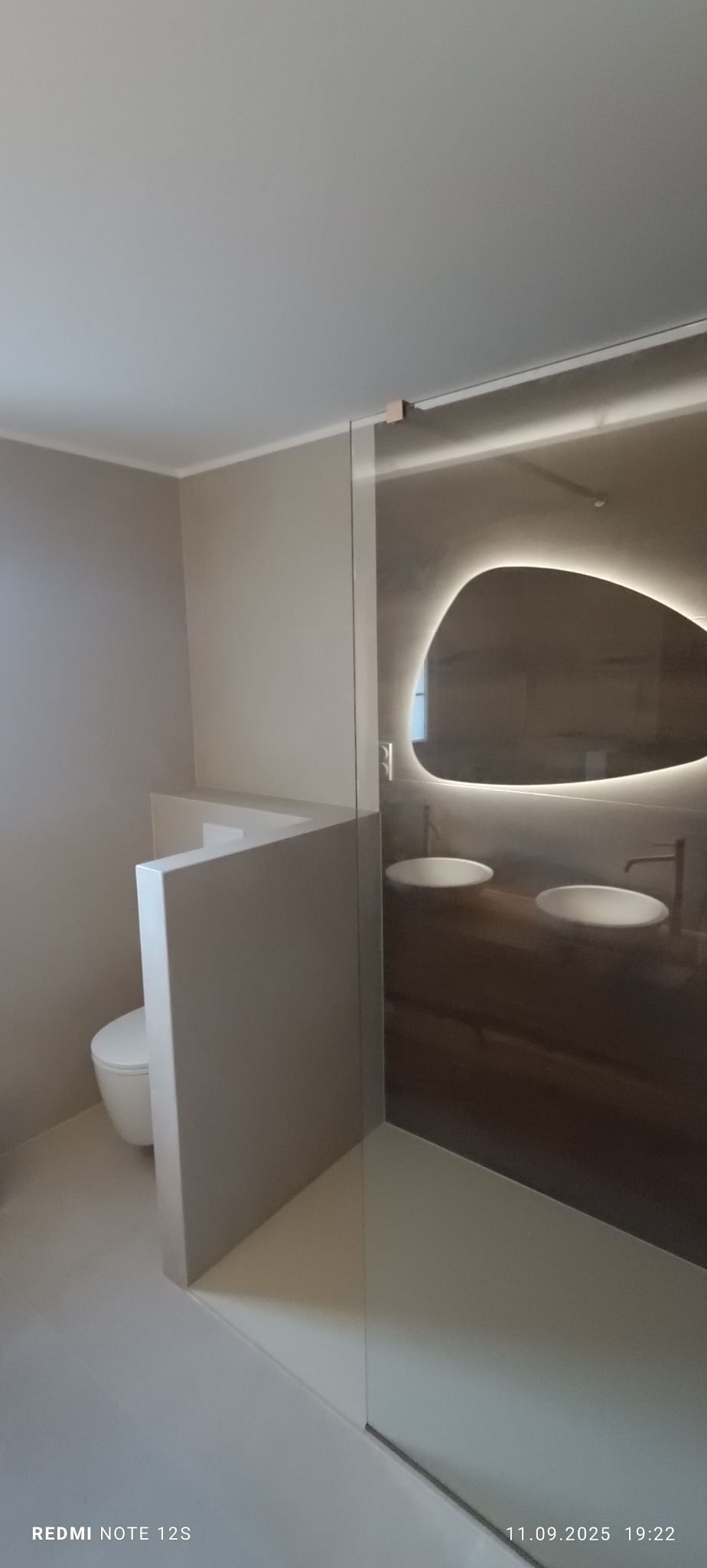 Nowoczesna łazienka w beżach z podświetlanym lustrem o nieregularnym kształcie, kabiną prysznicową ze szkła i zabudowaną toaletą. Minimalistyczny design.