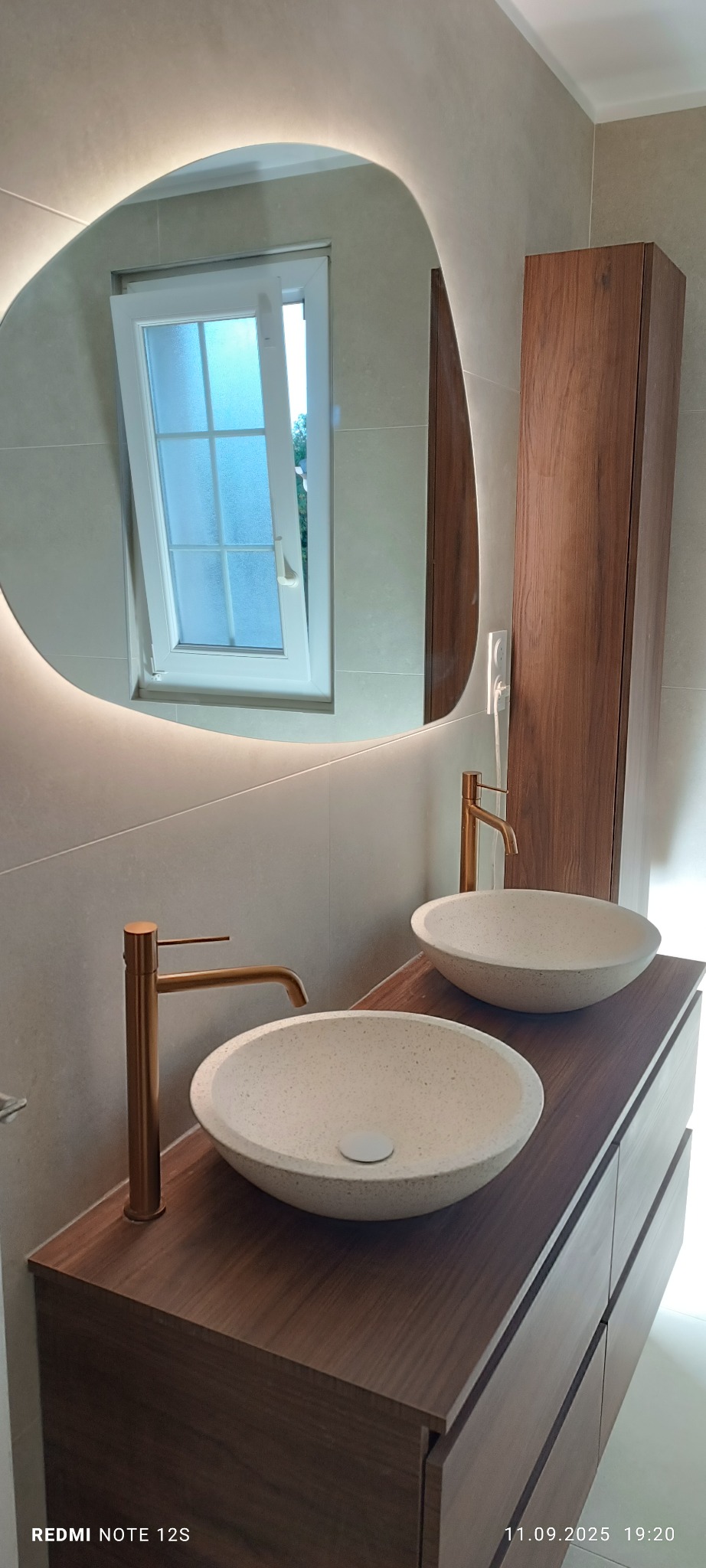 Nowoczesna łazienka z dwoma umywalkami nablatowymi, złotymi bateriami i podświetlanym lustrem o nieregularnym kształcie. Widoczne okno z otwartym skrzydłem.