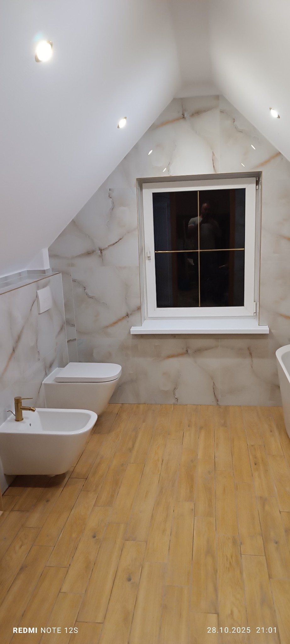 Jasna łazienka na poddaszu z marmurowymi płytkami ściennymi i drewnopodobną podłogą. Uwagę przykuwa złota armatura przy bidecie i odbicie osoby w oknie.