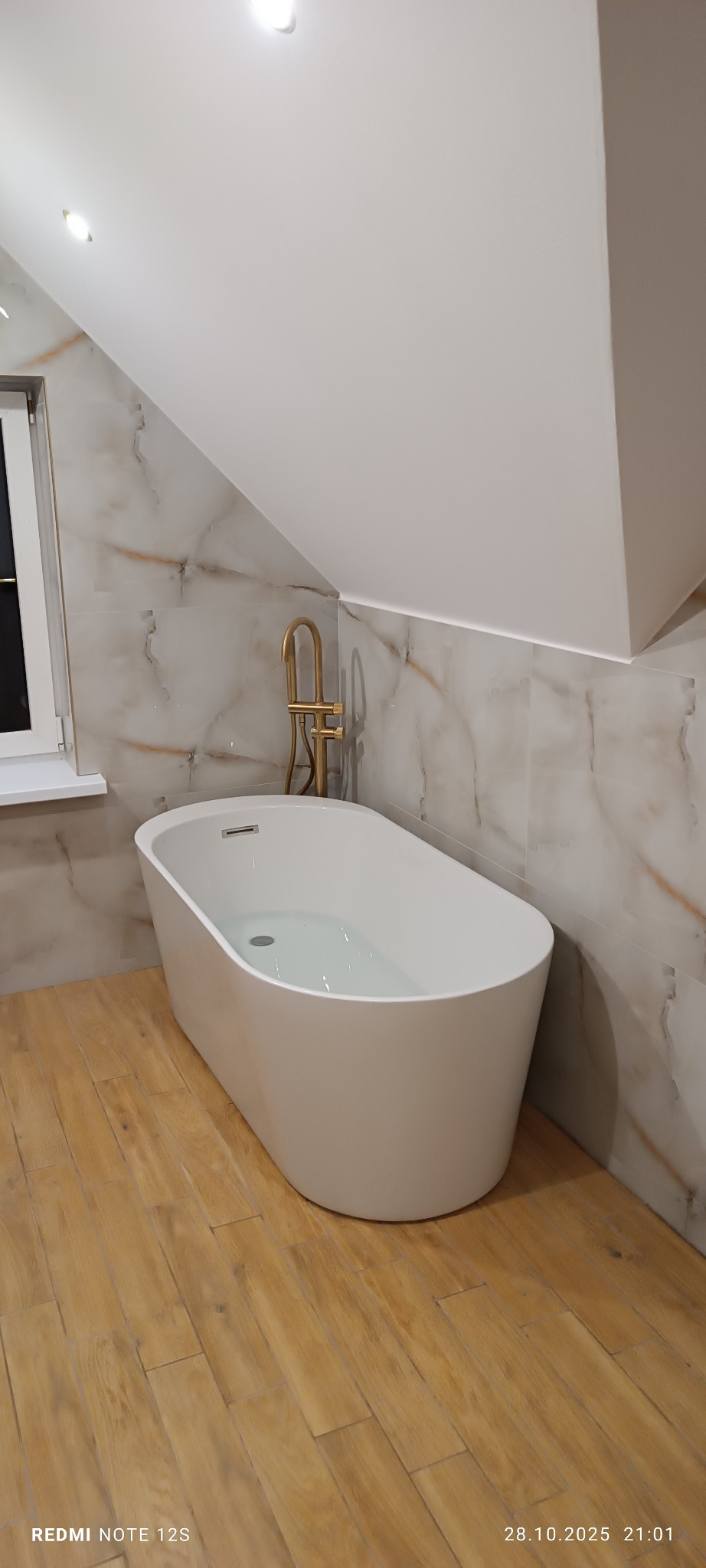Nowoczesna, wolnostojąca wanna wypełniona wodą, z złotą armaturą na tle marmurowych płytek w łazience na poddaszu z drewnopodobną podłogą.