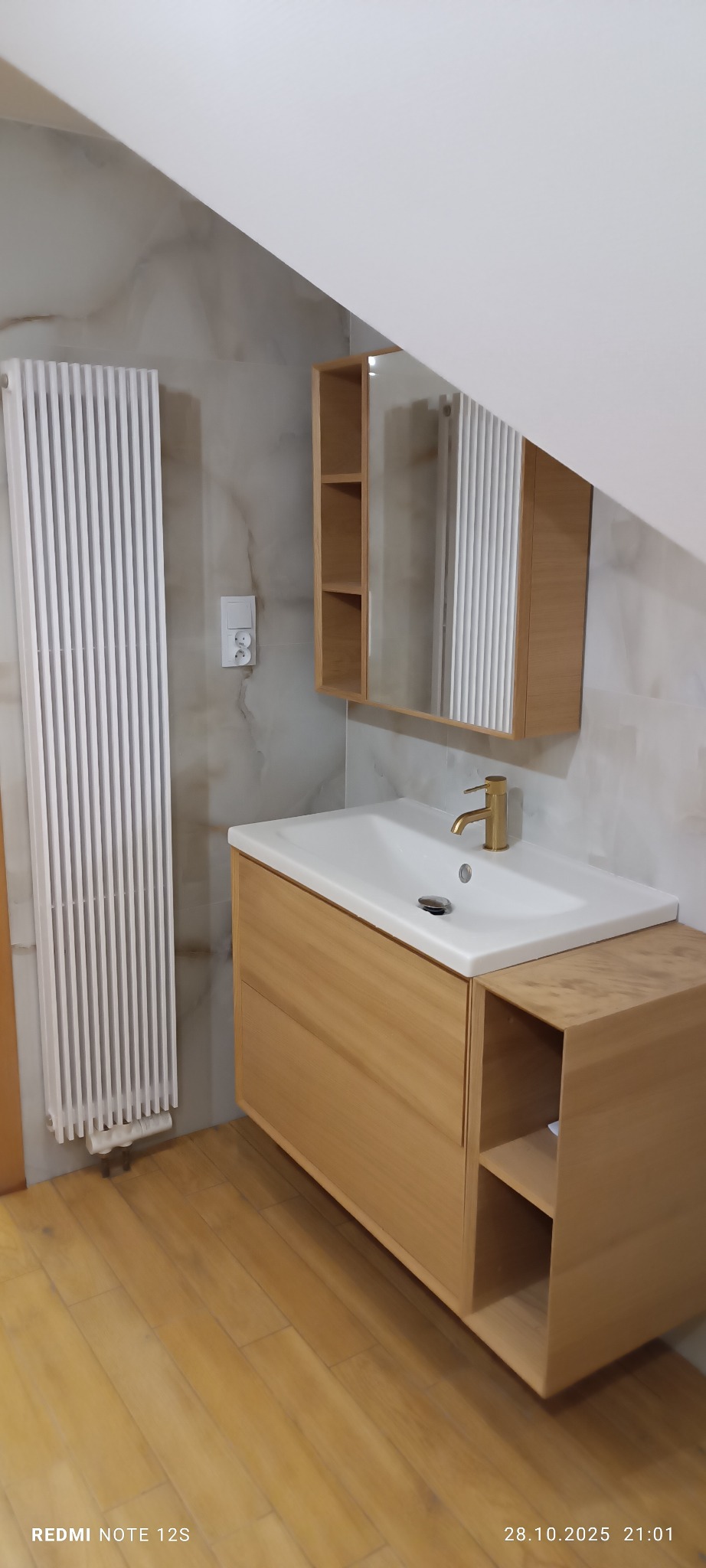 Jasna łazienka na poddaszu: drewniana szafka z umywalką, złota bateria, lustro z półkami, biały grzejnik żeberkowy, marmurowe płytki ścienne i drewniana podłoga.