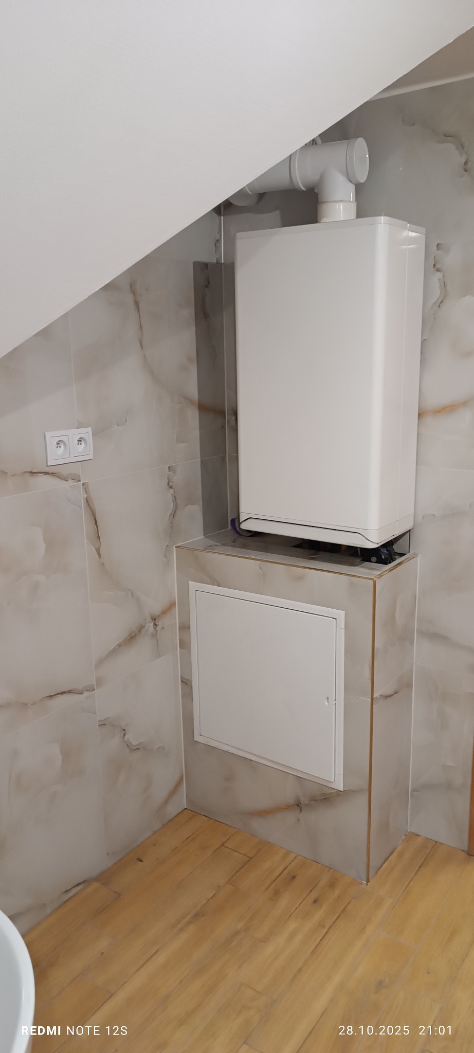 Biały piec gazowy z rurą odprowadzającą spaliny, zamontowany w łazience wykończonej marmurowymi płytkami i drewnopodobną podłogą. Widoczny fragment ściany ze gniazdkiem.