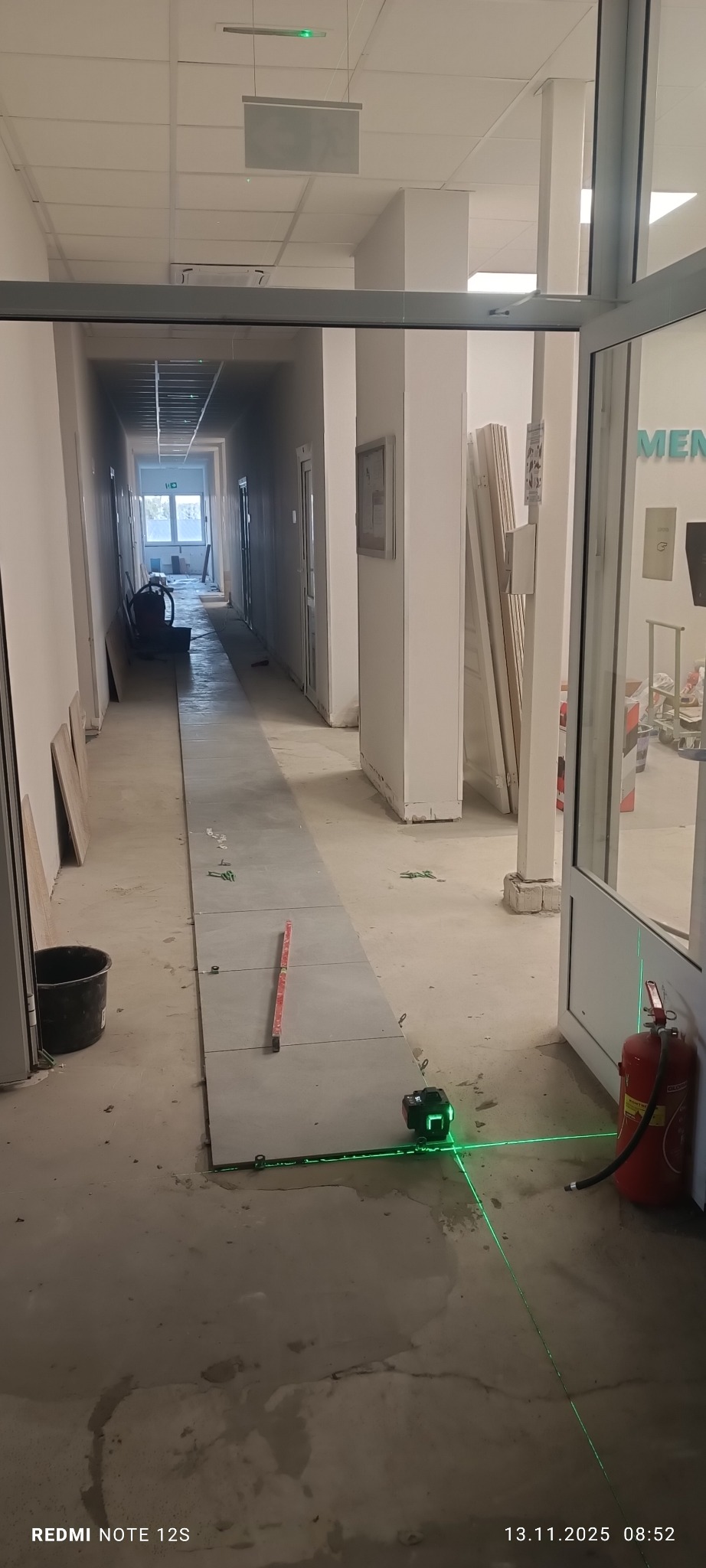 Kładzenie szarych płytek w długim korytarzu, poziomowanie laserem krzyżowym. Widoczny kubeł, poziomica i zapasowe płytki.