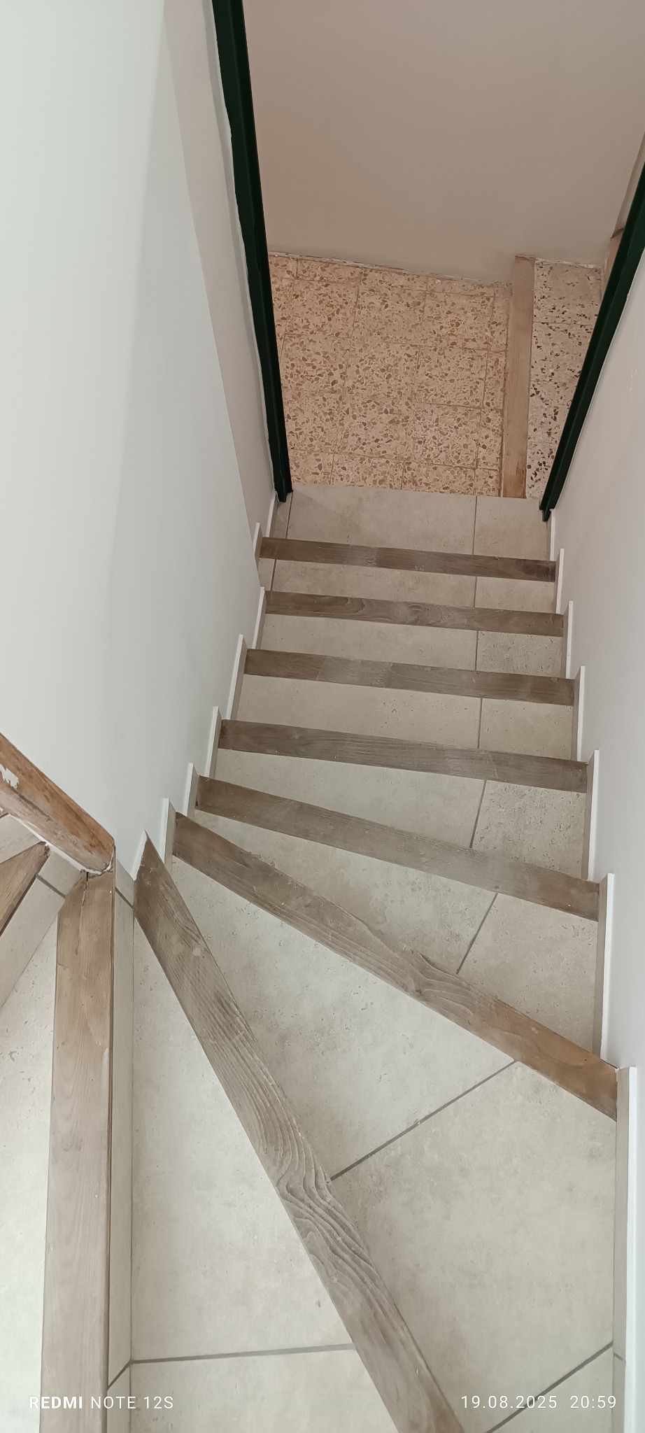 Wykończone schody z jasnymi płytkami i drewnianymi elementami, widok z góry. Białe ściany i ciemnozielone wykończenie krawędzi. Jasne, naturalne światło.