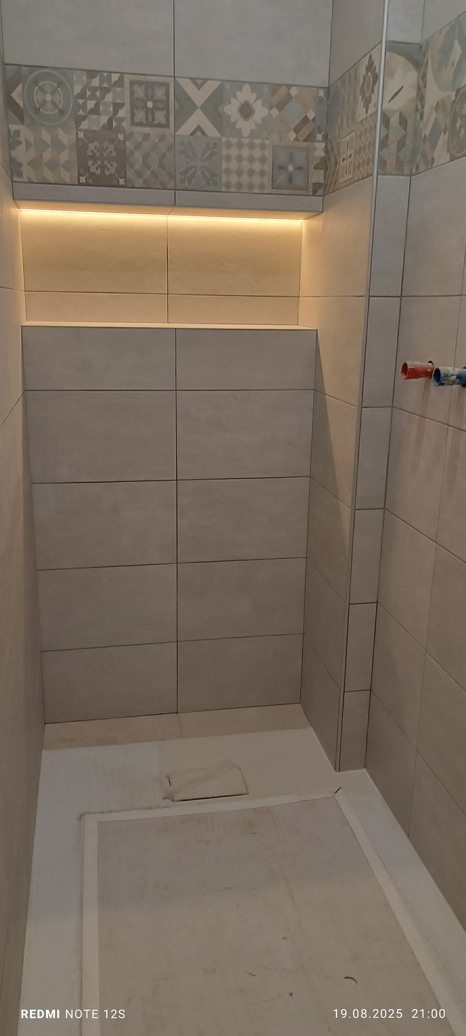 Wykończona kabina prysznicowa z beżowymi płytkami, dekoracyjnym pasem mozaiki i podświetlaną wnęką. Widoczne przyłącza wodne i brodzik w trakcie przygotowania.