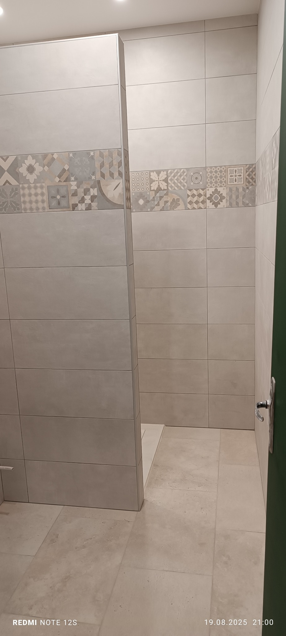 Wykończona łazienka z jasnymi, poziomymi płytkami i dekoracyjnym pasem z wzorami. Widoczny fragment kabiny prysznicowej i podłoga z płytek w odcieniach beżu.